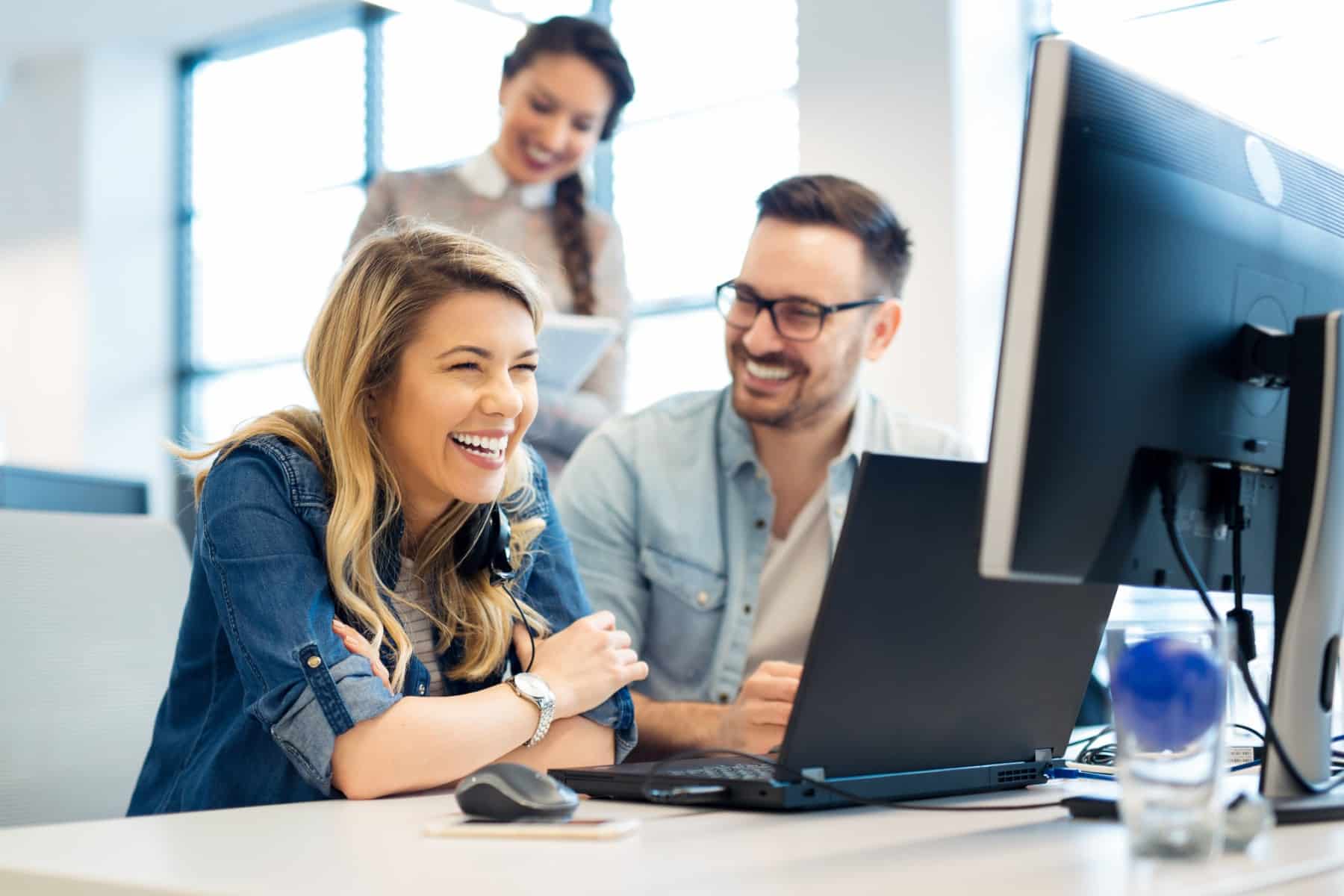 Three people smiling and working together at computers in a bright office.