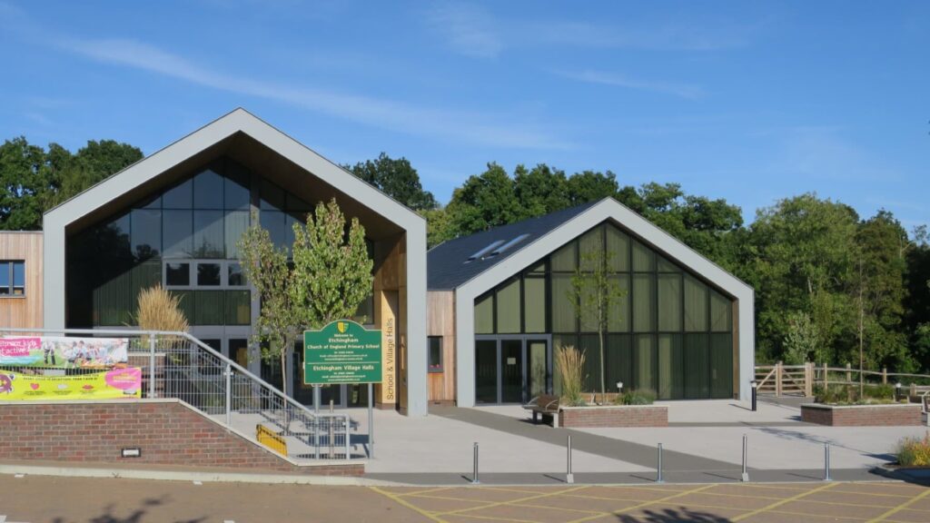 Modern school building with large windows, green sign, and trees in front under a clear blue sky.