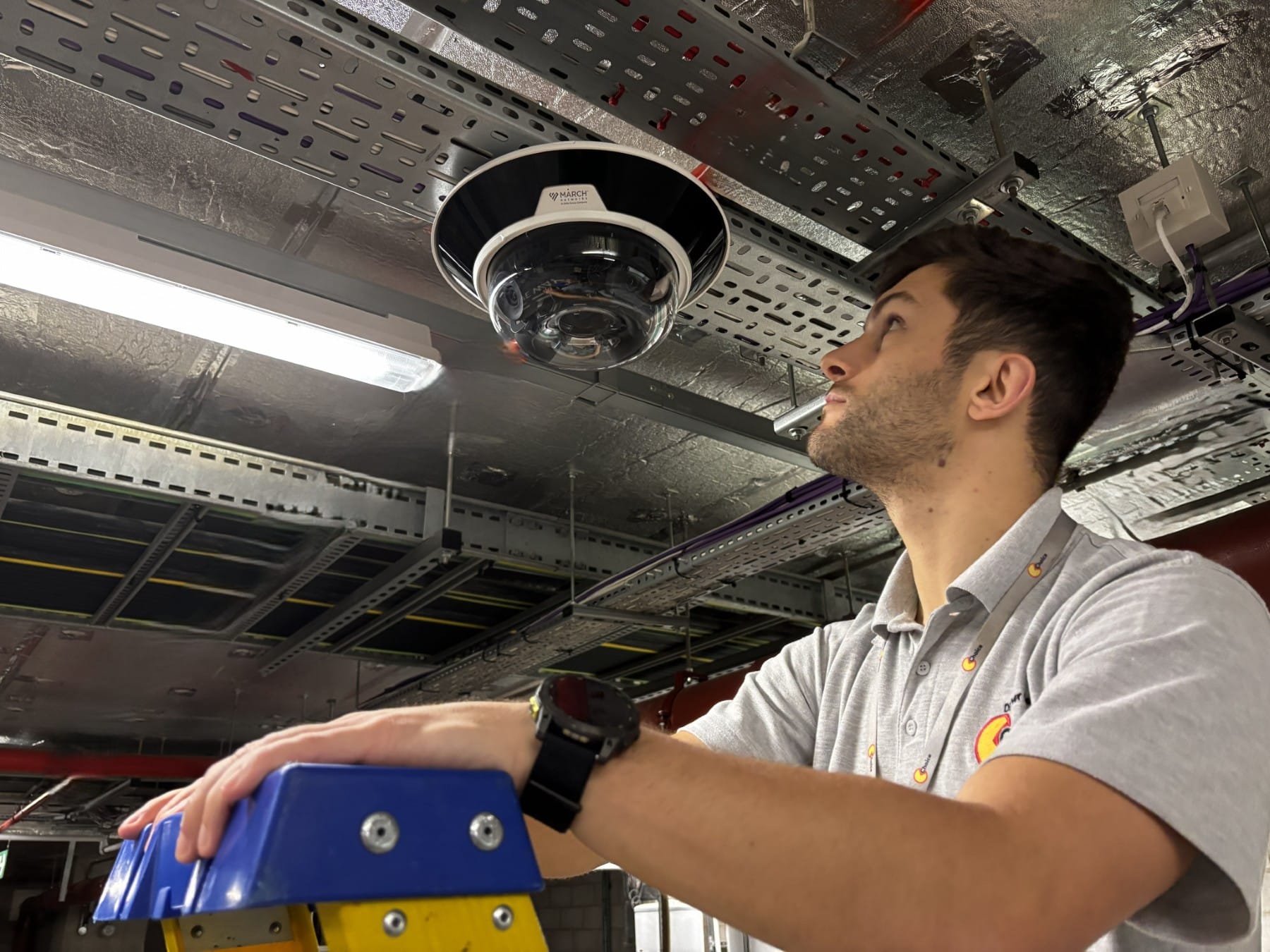 Man on ladder installing a CCTV camera on a ceiling in an industrial building.