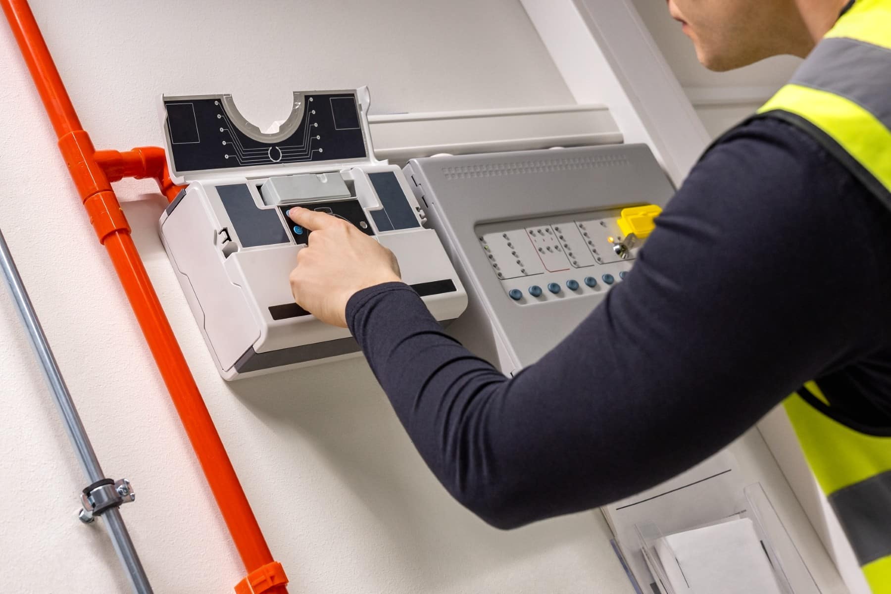 Technician testing a fire alarm control panel on a wall with red piping.