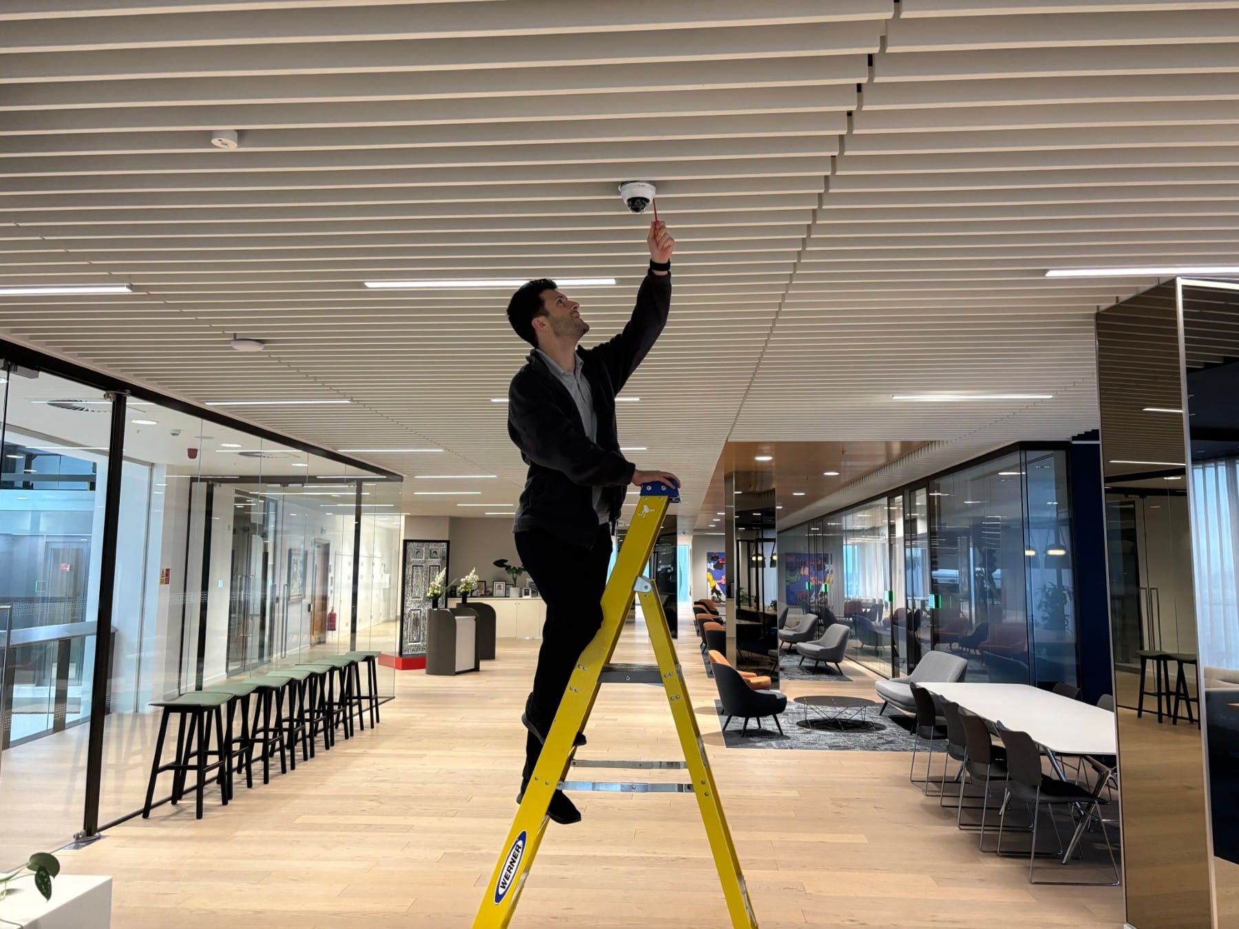 A man stands on a yellow ladder installing or adjusting a ceiling camera in a modern office space.