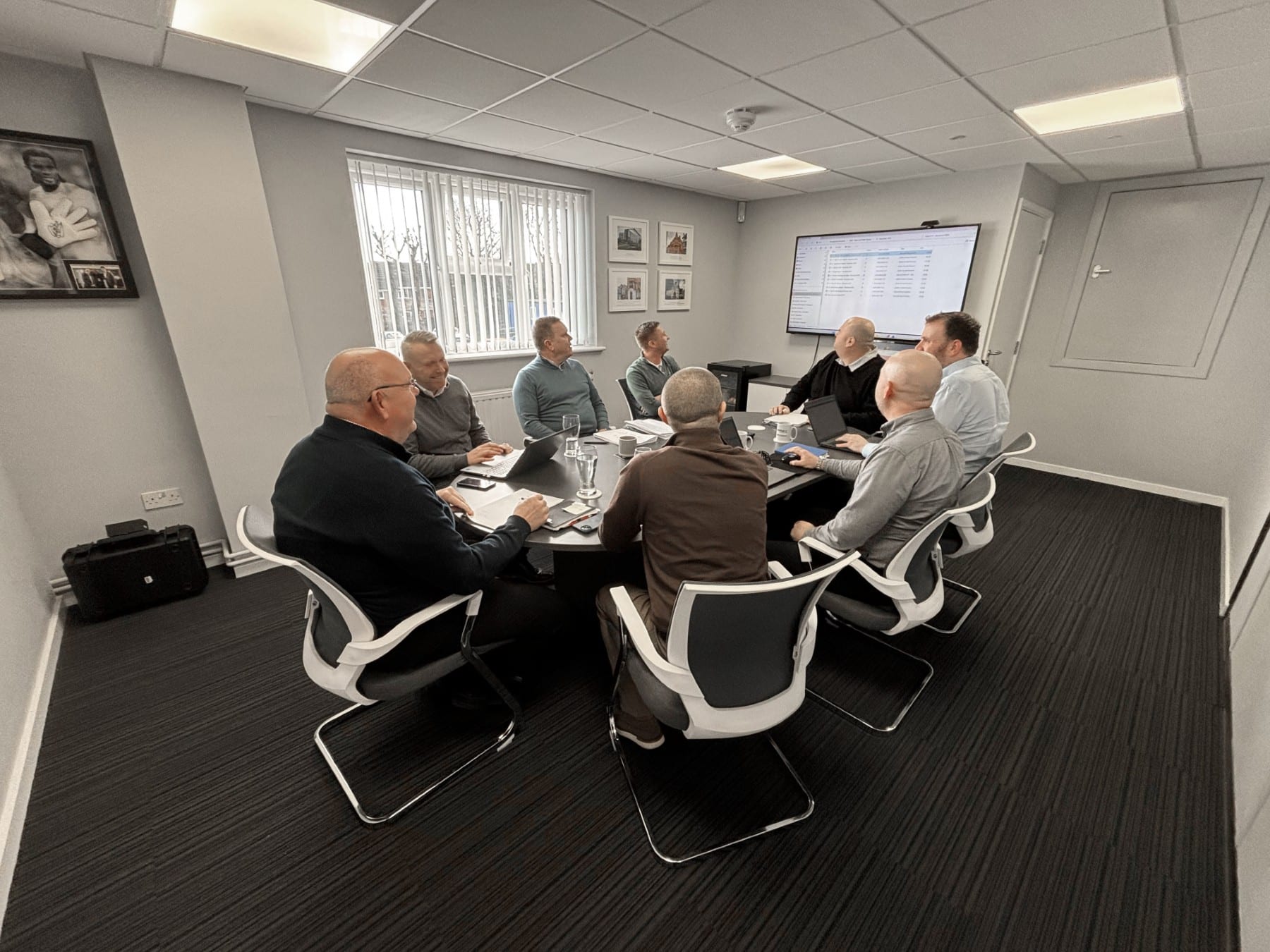 Eight people sit around a table in a meeting room, looking at a screen on the wall.