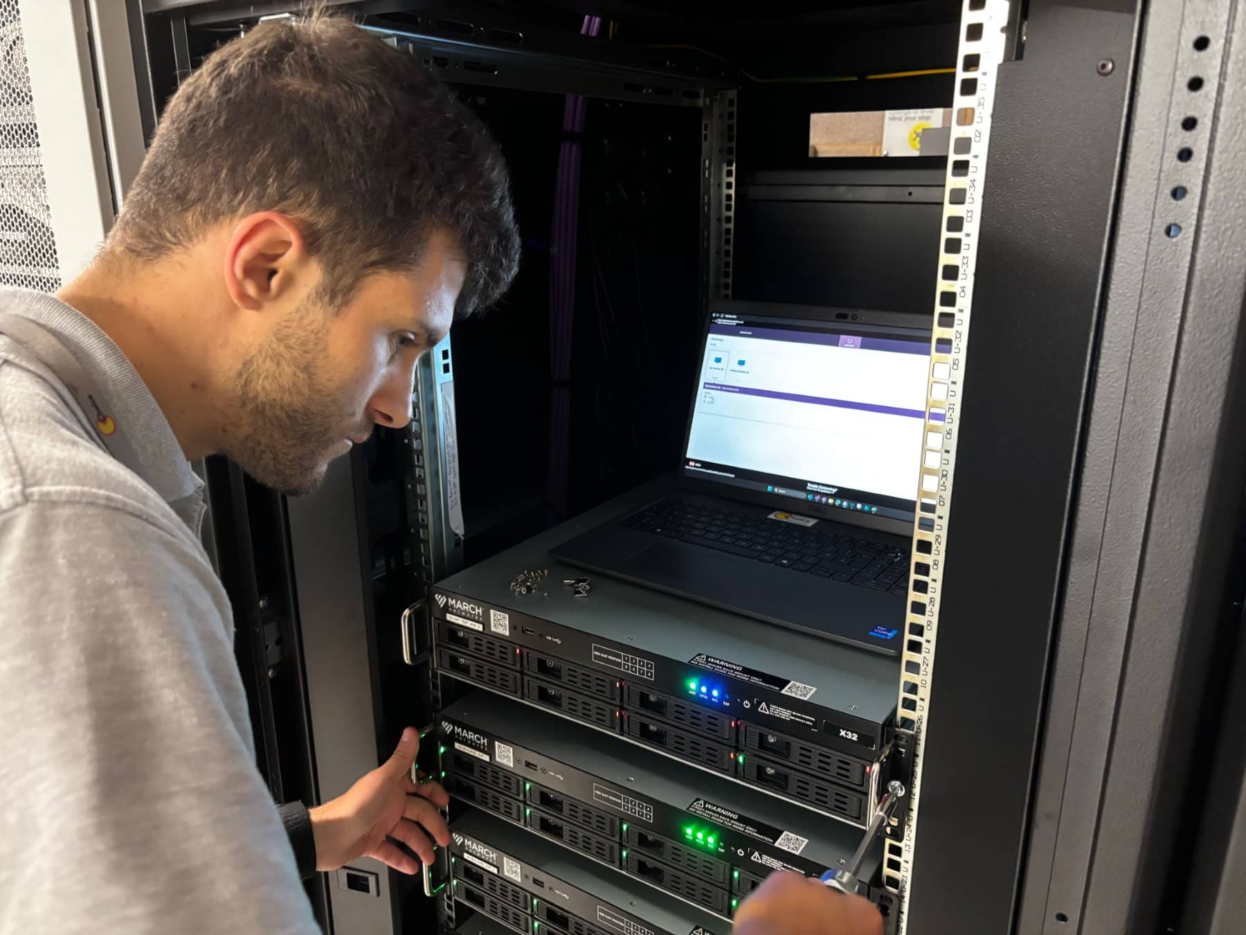 Person working with computer servers in a server rack, with a laptop open above the servers.