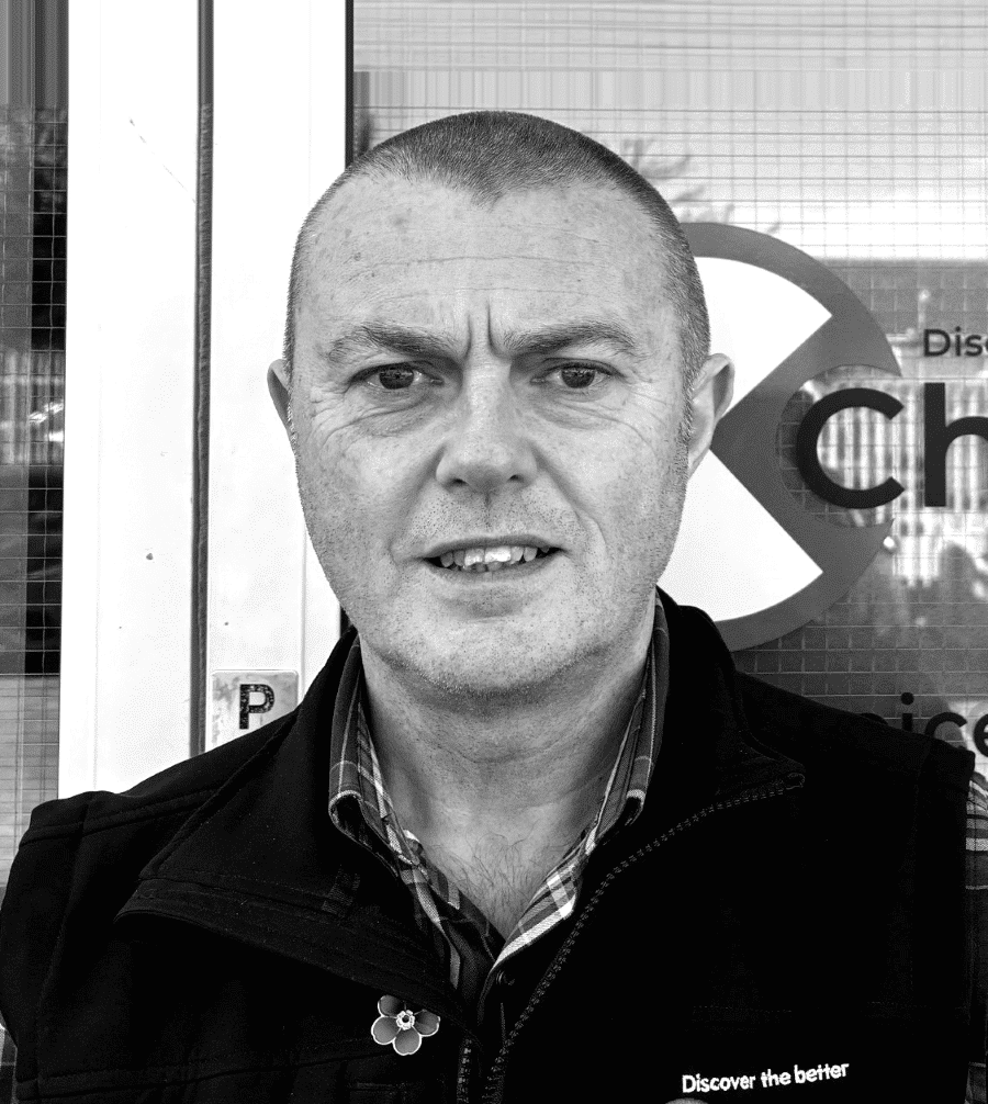 A man with short hair stands outside, wearing a jacket and shirt, looking at the camera.
