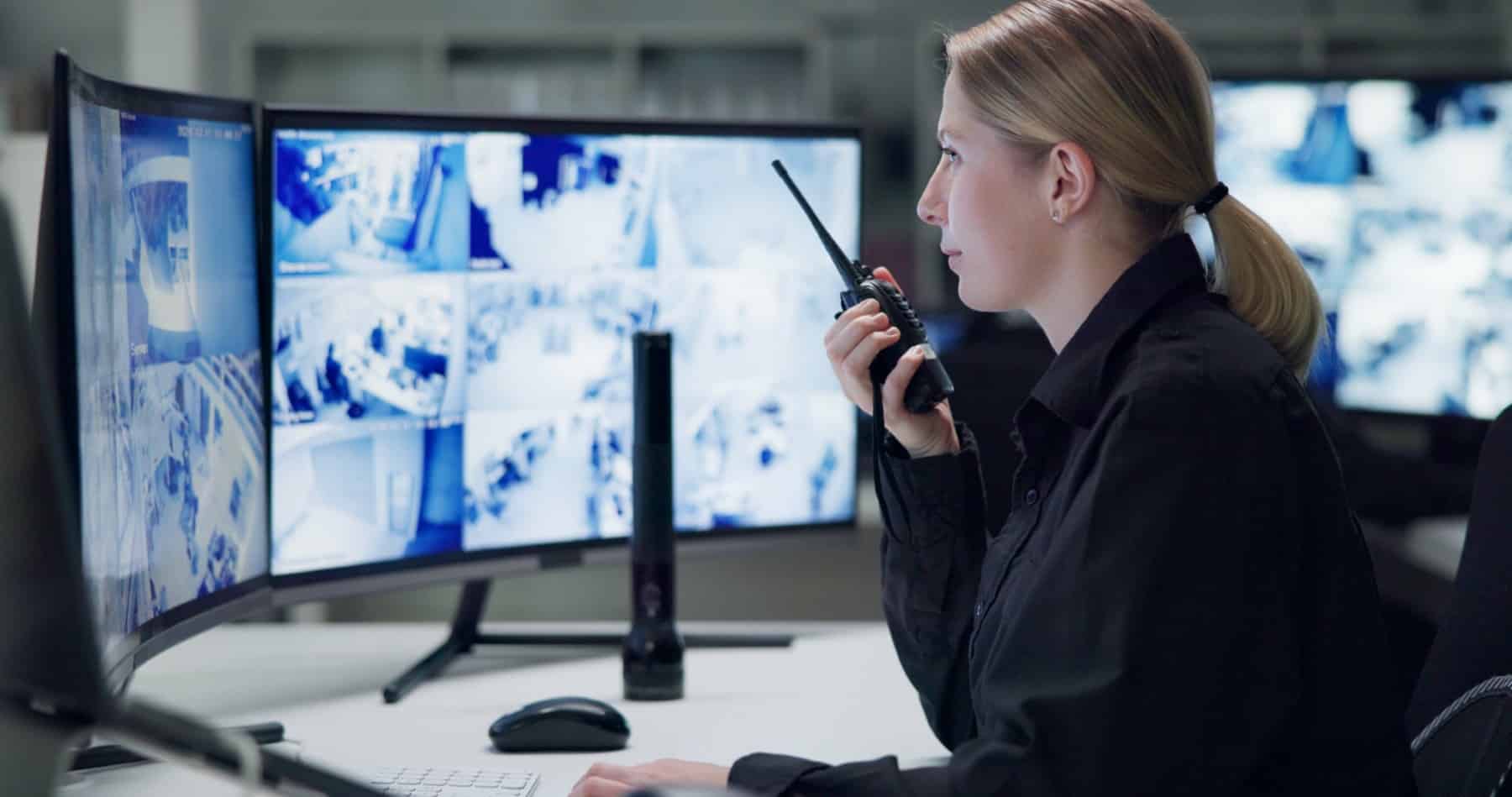 A woman monitors security cameras on multiple screens and speaks into a walkie-talkie.