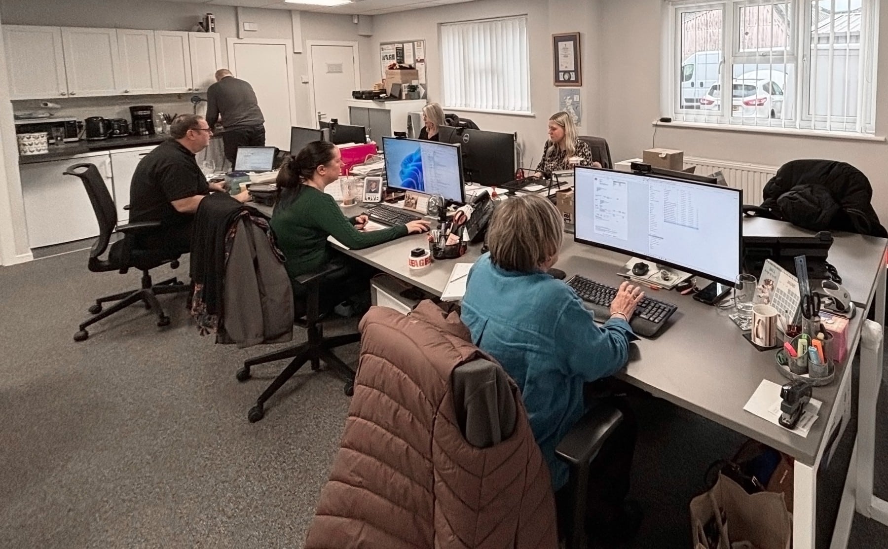 Office space with people working at desks and computers, one person in the kitchen area in the background.