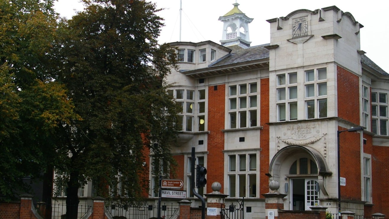 Historic brick building with large windows and a white entrance, partially obscured by trees.