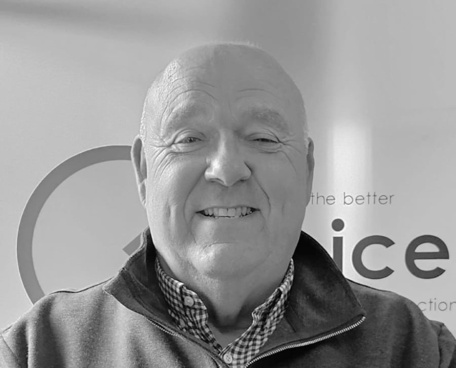 Smiling older man in a collared shirt and zip-up sweater, in front of a sign. Black and white photo.