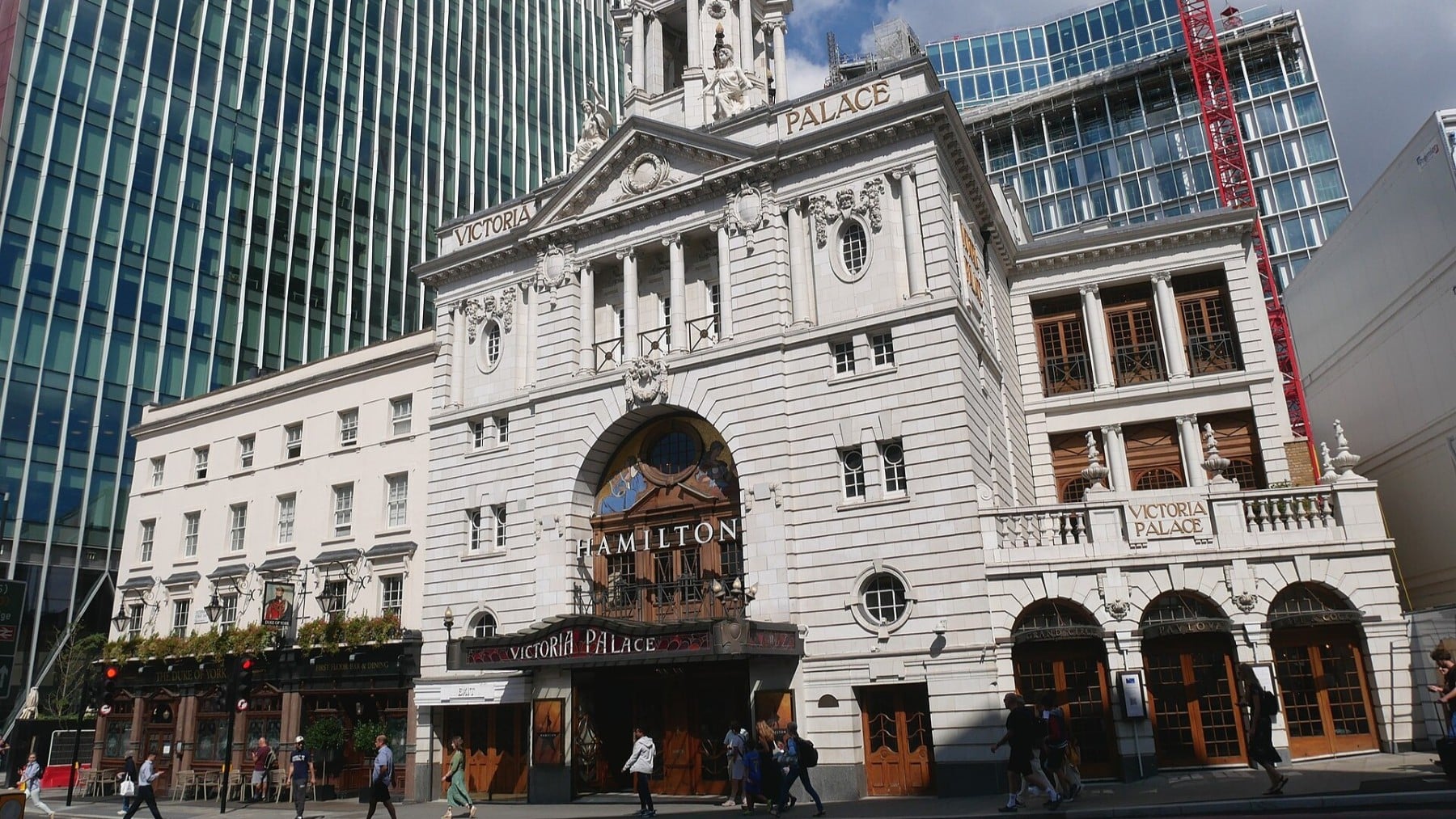 Victoria Palace Theatre with people walking outside, surrounded by modern buildings.