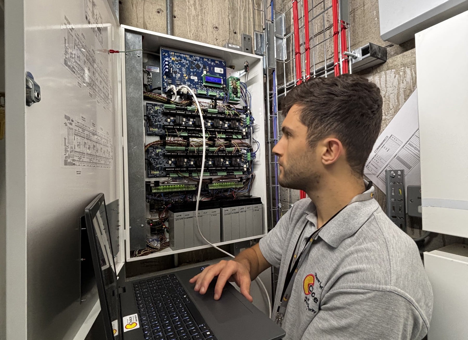 Our Partners - See Our Projects Man working on a laptop next to an open electrical control panel in a building.